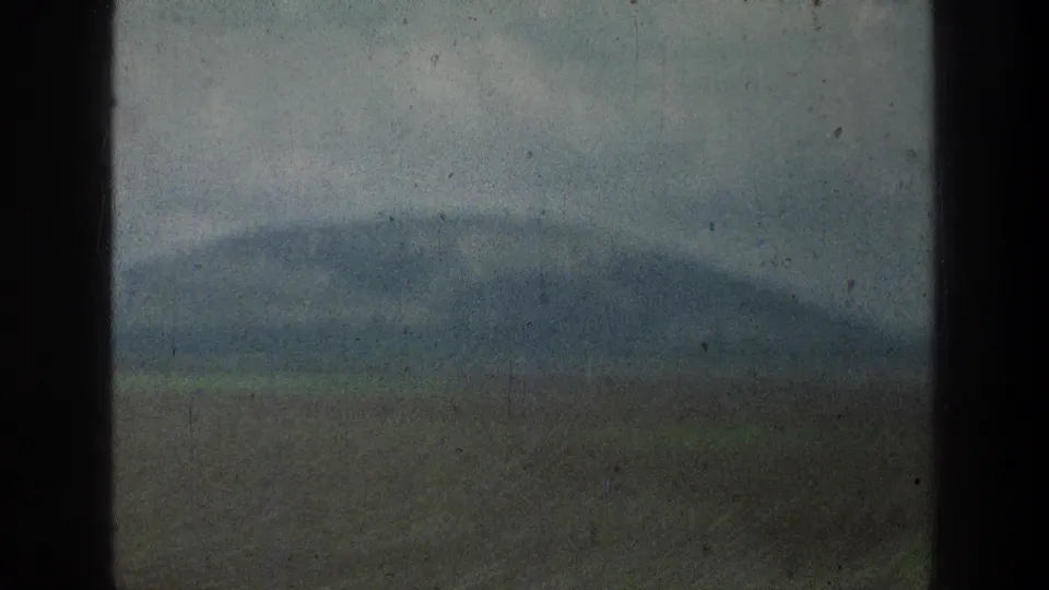 A muted 8mm landscape frame shows distant hills, open ground, and cloudy sky through a window in the cataloged 1976 Jacobs Well Jerusalem clip.