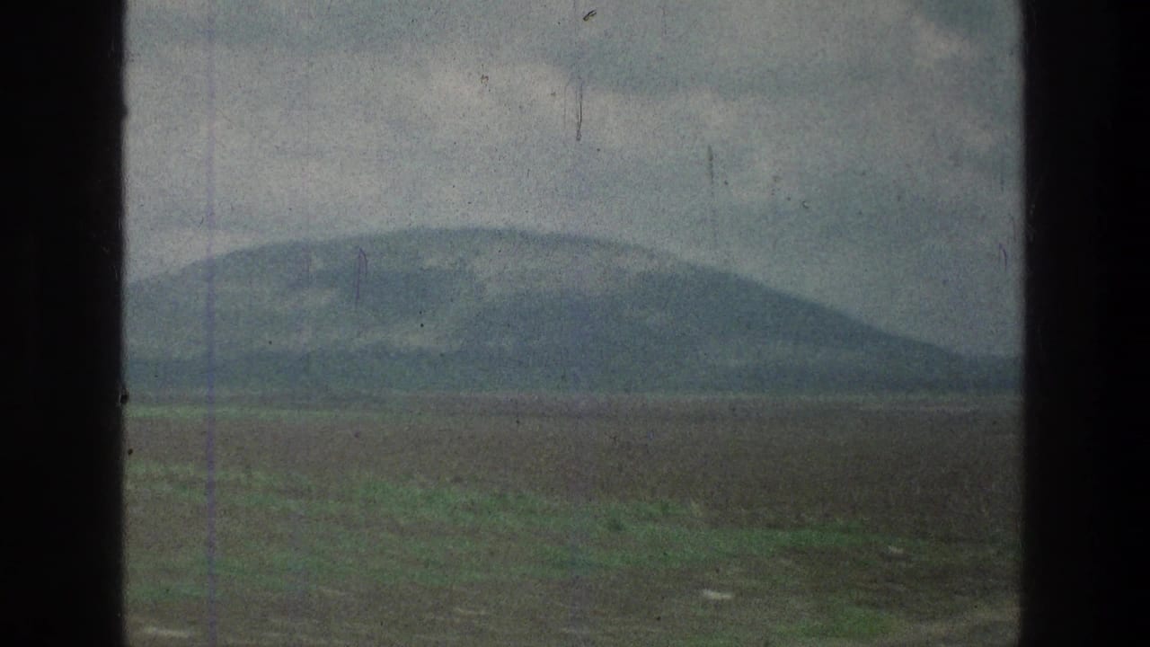Moving 8mm view of a low mountain and broad field, partially blocked by dark vertical edges in the frame.