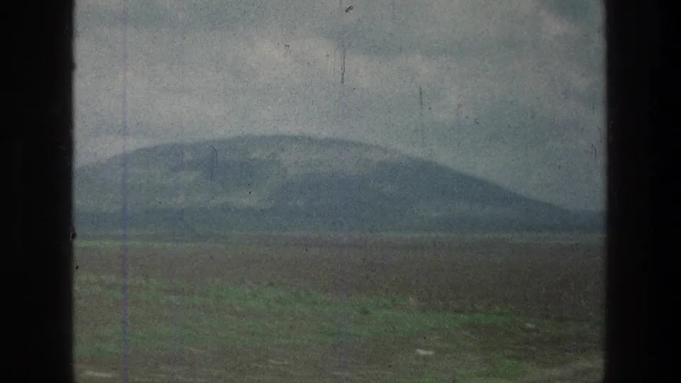 Moving 8mm view of a low mountain and broad field, partially blocked by dark vertical edges in the frame.