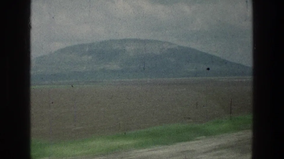1976 home-movie frame of fields and a low hill, with a green roadside strip crossing the foreground.