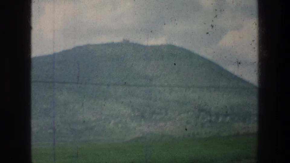 Moving-vehicle view from 1976 8mm footage showing a rounded green hill, cloudy sky, and a horizontal power line near the stated Jerusalem-area location.