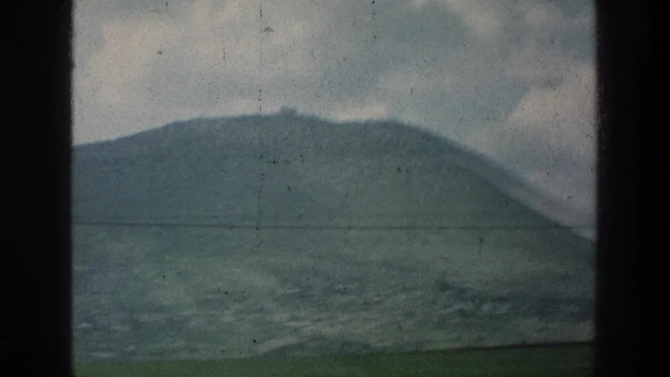 1976 8mm travel footage frame showing a soft green hill and lower terrain through dark frame borders, with visible grain and scratches.