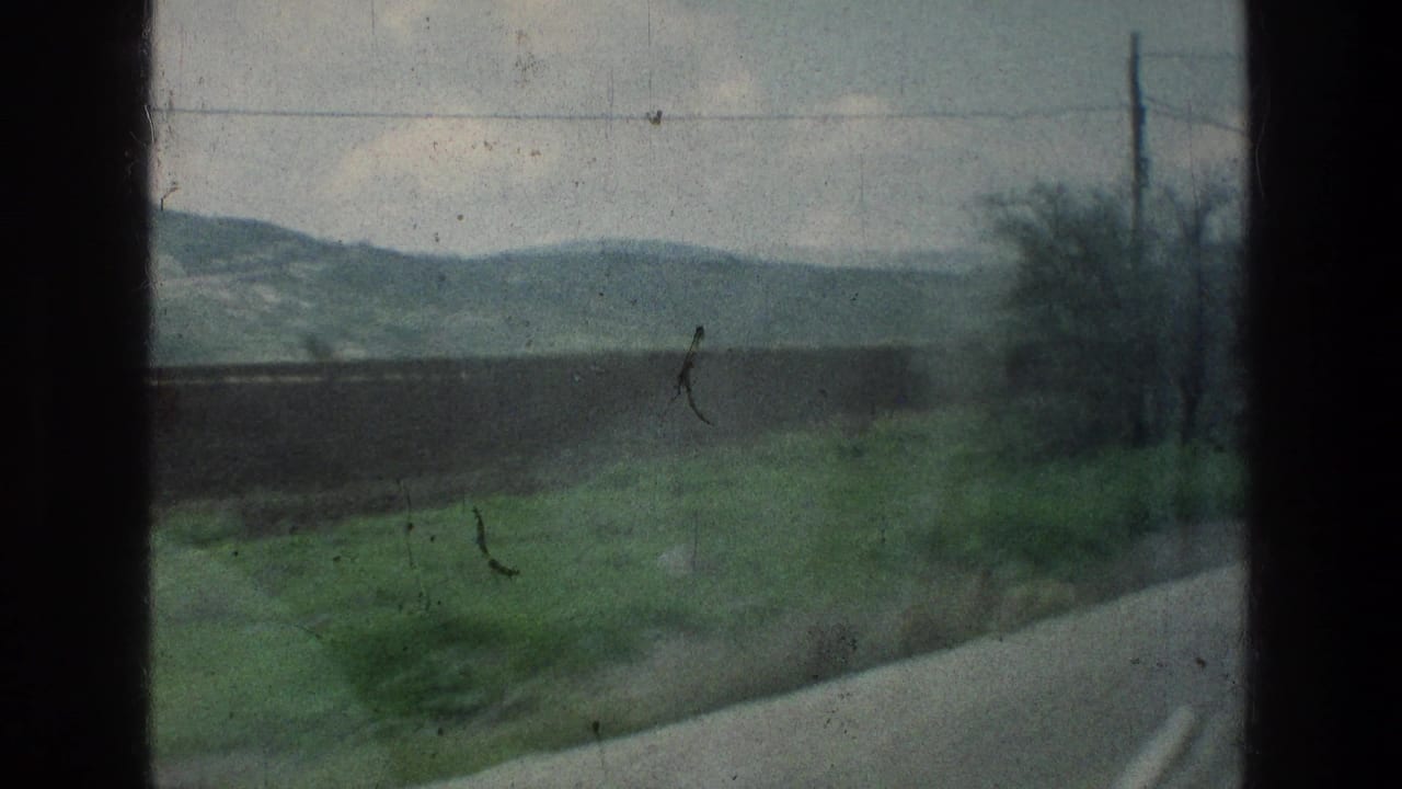 1976 8mm travel view from a moving vehicle near the stated Jacobs Well Jerusalem location, showing road edge, fields, brush, hills, and cloudy sky.