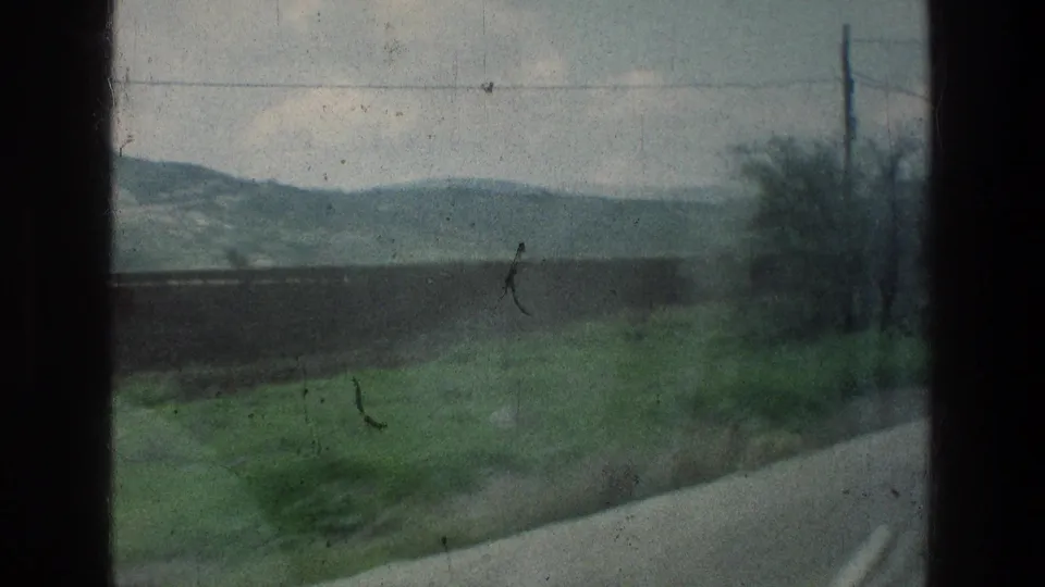 1976 8mm travel view from a moving vehicle near the stated Jacobs Well Jerusalem location, showing road edge, fields, brush, hills, and cloudy sky.