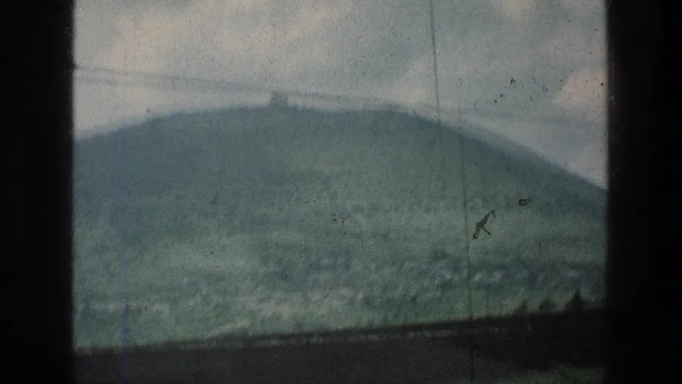 1976 8mm frame showing a distant green hill and small ridge-top structure in the stated Jacobs Well Jerusalem area, viewed through scratched film.