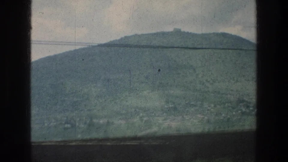 Final 8mm travel frame of a green hill in the stated Jerusalem-area setting, with a small ridge-top structure, wire line, and cloudy sky.