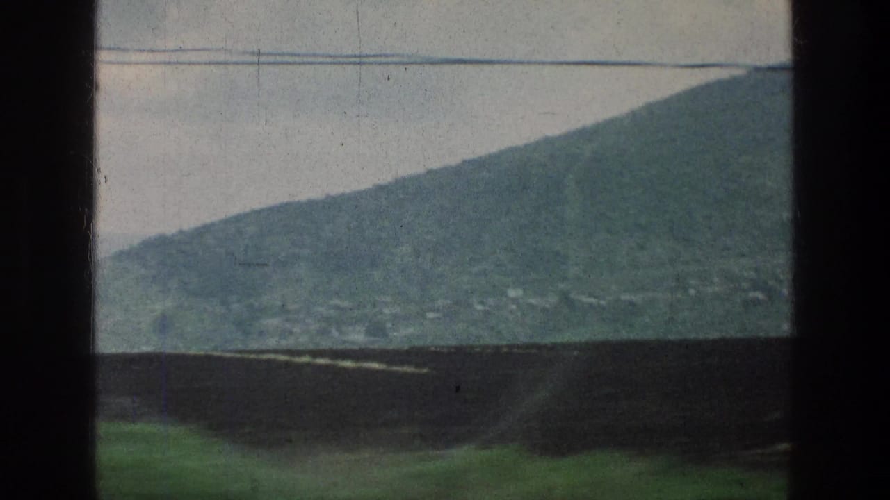 Rounded green hill, pale sky, power lines, and roadside wall viewed from a vehicle.