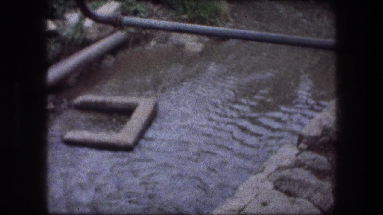Archival 8mm frame of a rippled gray water channel in Jericho with a pipe and concrete piece visible.