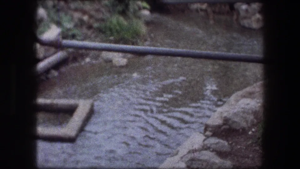 Jericho home-movie frame showing a murky stream crossed by a pipe with rocks and concrete beside it.