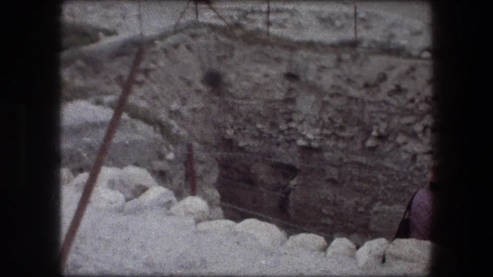 Final 1976 Jericho frame showing the rocky excavation face, pale foreground stones, and a partially visible visitor at the right edge.
