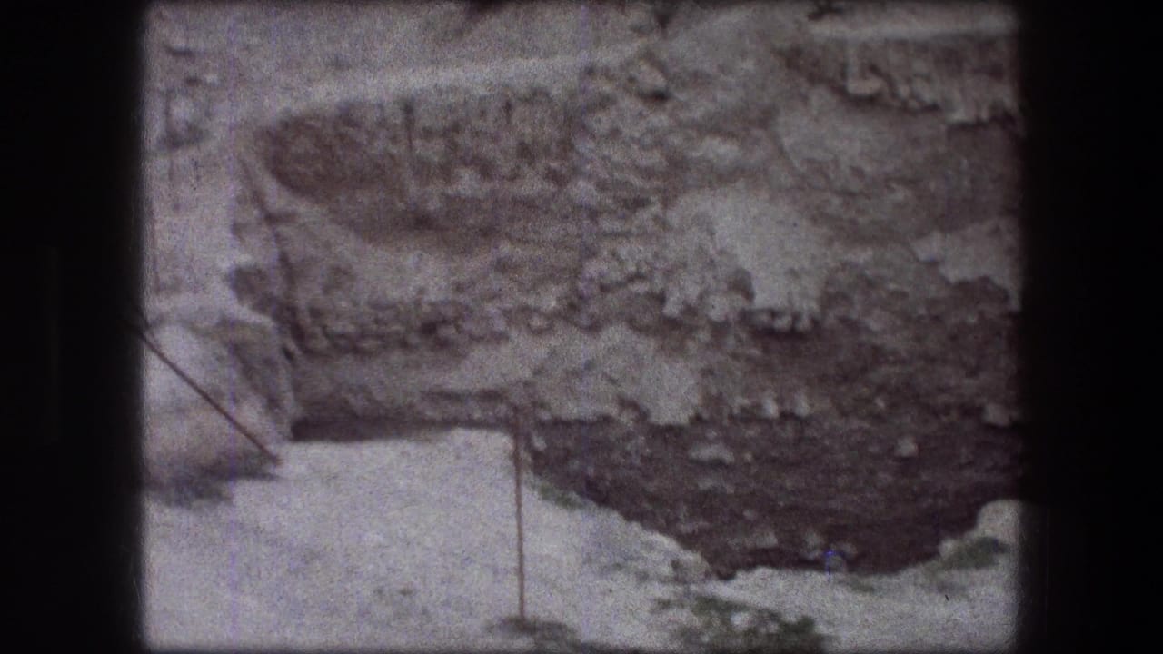 1976 Jericho frame with a textured rock face, pale ledge, sparse greenery, and a thin metal post marking the edge.