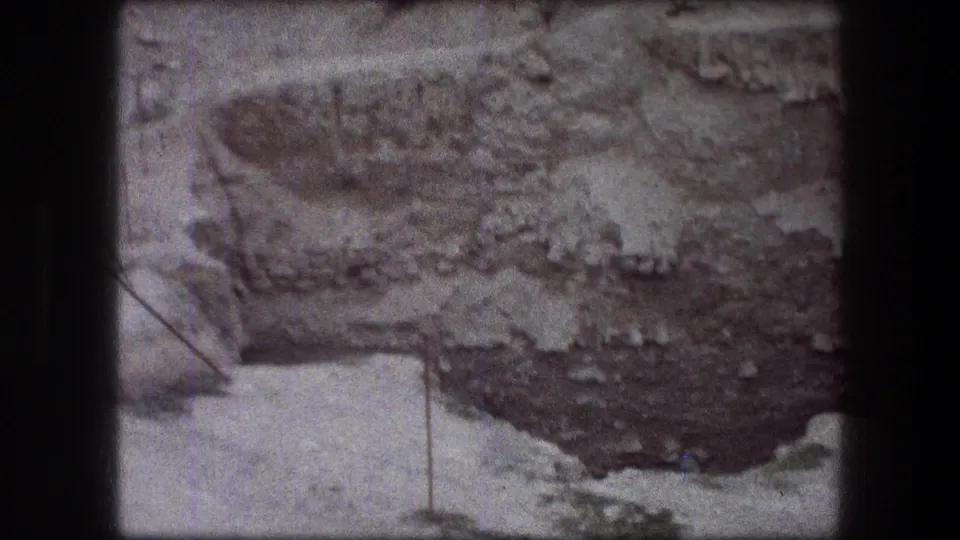1976 Jericho frame with a textured rock face, pale ledge, sparse greenery, and a thin metal post marking the edge.