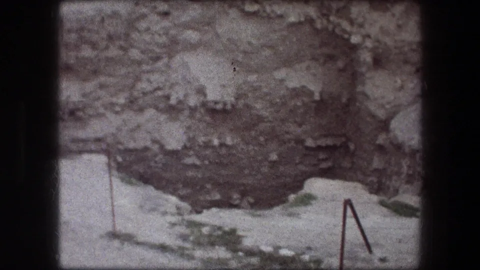 1976 Jericho 8mm frame showing pale ground before a dark rocky recess, with a small angled metal railing near the lower edge.