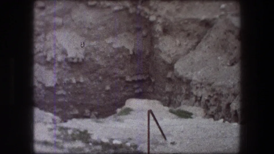 1976 Jericho frame showing a weathered rock or wall surface, pale ledge, scattered greenery, and an angled metal railing.