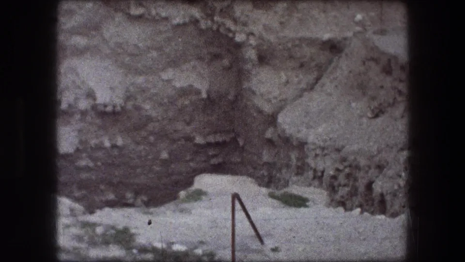 1976 Jericho 8mm frame showing rough excavated earth, pale gravel, stone layers, and a thin metal stake in the foreground.