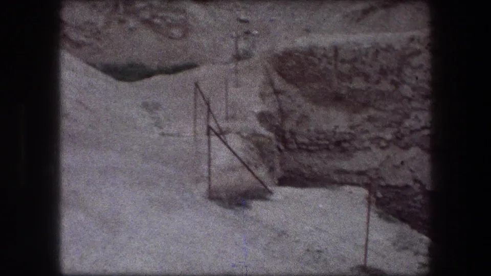 1976 Jericho 8mm frame showing metal stakes near a gravel edge, rough excavated wall, and pale disturbed ground.