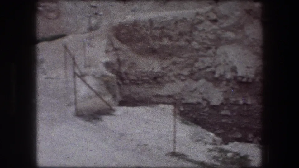 1976 8mm frame from Jericho showing a rough rocky pit edge, pale earth, sparse vegetation, and simple metal boundary posts.