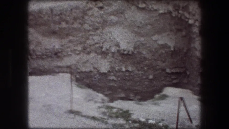 Jericho, Israel home-movie frame showing layered rock, pale terrain, small green patches, and a simple metal railing beside the pit.