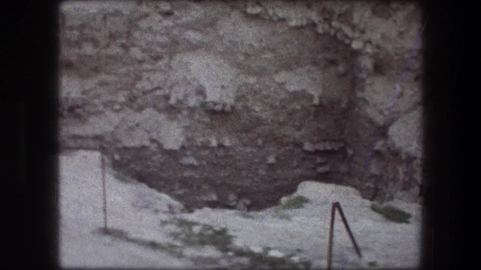 1976 8mm view of the Jericho pit area, with rough rock, pale soil, metal boundary posts, and scattered low vegetation.