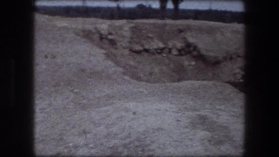 Wider Jericho frame showing pale uneven ground, a rocky depression, low metal railing, and a distant dark horizon under daylight.
