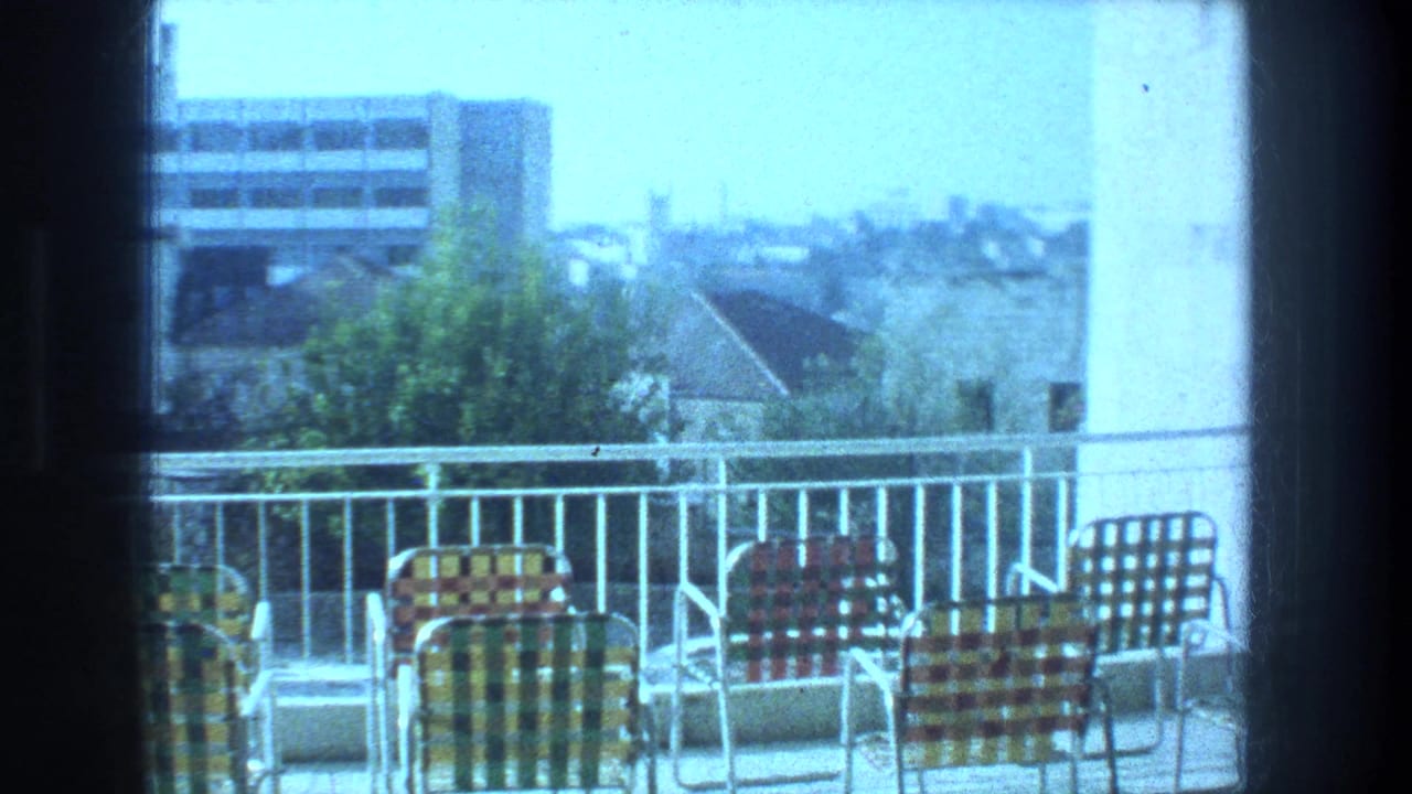 1976 8mm Jerusalem rooftop view with checkered patio chairs, white railing, trees, rooftops, and a hazy skyline.