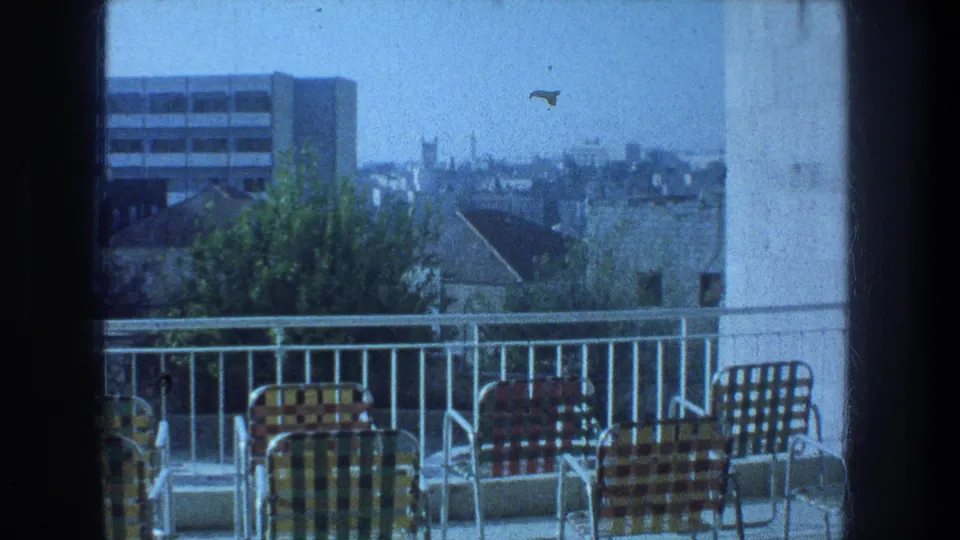 Jerusalem rooftop balcony scene with colorful checkered chairs, a white rail, trees, and distant urban buildings on 8mm film.