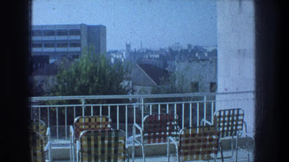 1976 Jerusalem balcony view showing checkered metal chairs, a white railing, nearby trees, rooftops, and a modern building in the distance.