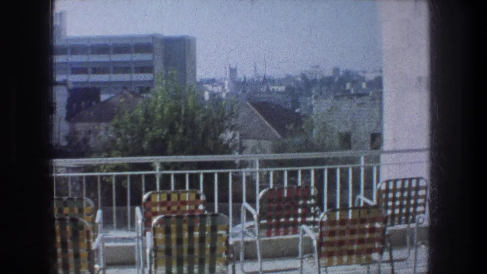 Jerusalem rooftop balcony in 1976 with patterned patio chairs, white railing, greenery, rooftops, and a soft distant skyline.