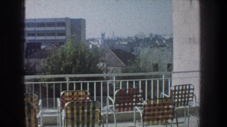 1976 Jerusalem balcony view with patterned lawn chairs, white railing, nearby rooftops, trees, and a hazy urban skyline.