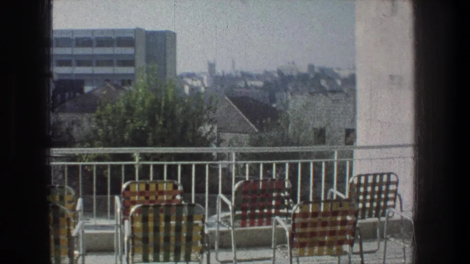 1976 Jerusalem home-movie frame showing colorful lawn chairs on a balcony with rooftops, trees, and a distant modern building.