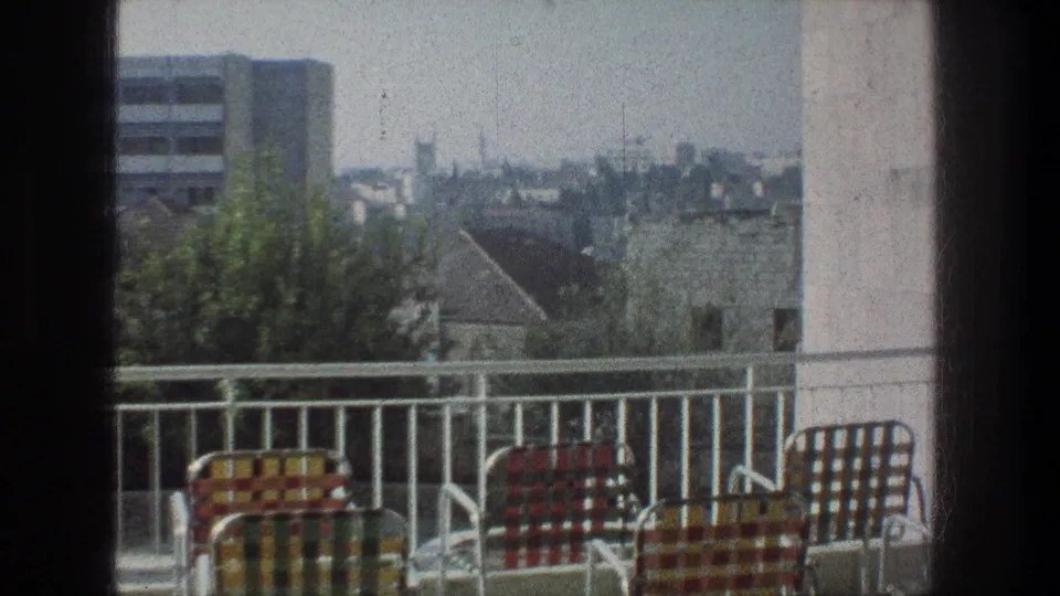 Jerusalem balcony view from 1976 with cropped lawn chairs, a white railing, rooftops, trees, and a soft distant skyline.