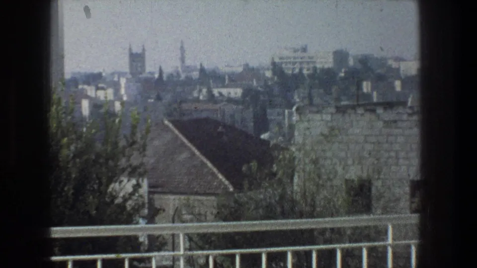 1976 8mm view from height over Jerusalem, showing rooftops, stone buildings, trees, and a balcony rail under hazy light.