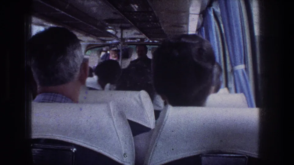 Interior of a Jerusalem bus in 1976, viewed from behind seated passengers with white seat backs, blue curtains, and overhead compartments.