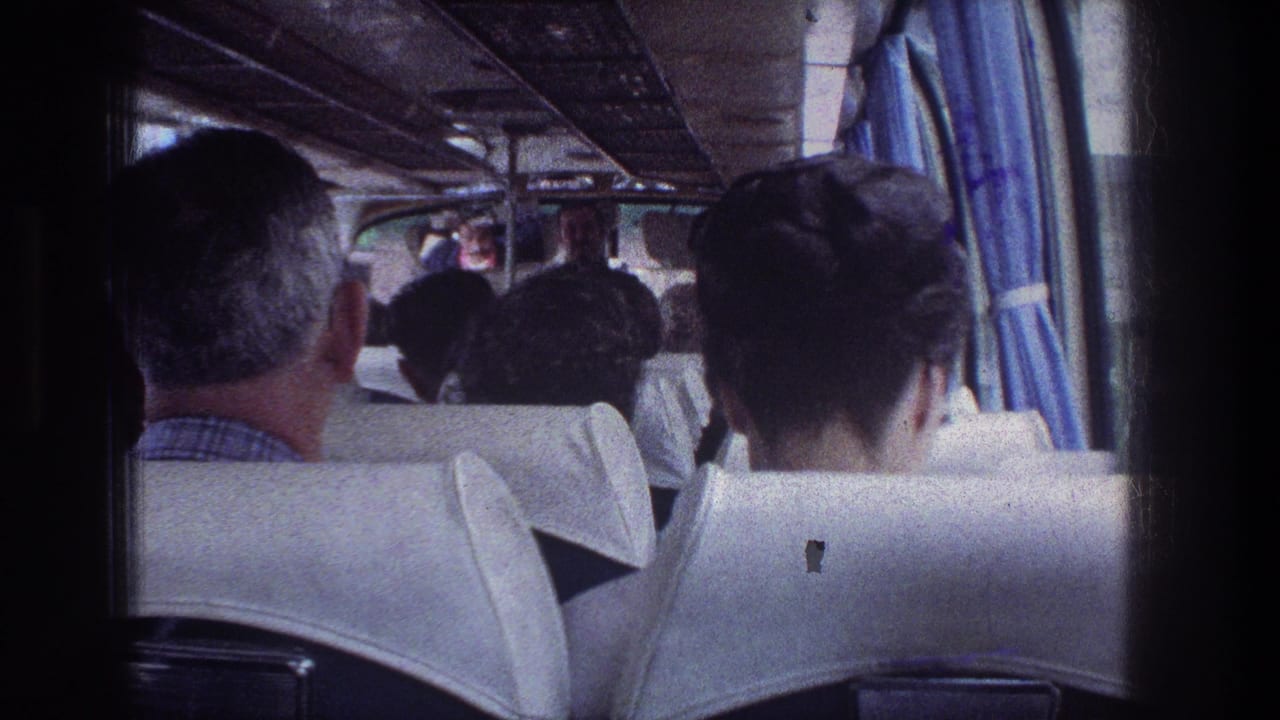 Passengers seen from the rear of a bus with white seat backs and blue curtains.