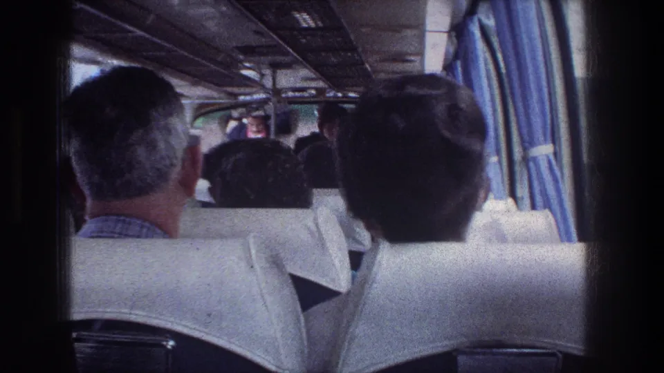 Inside the Jerusalem bus, adult passengers face forward while the handheld camera looks past white seats and blue curtains toward the front aisle.