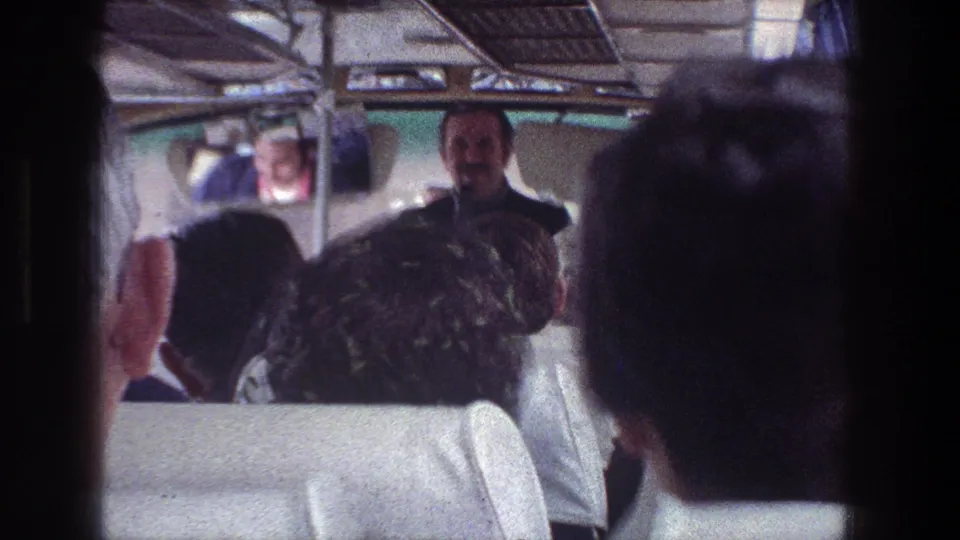 From the rear of the bus, passengers are seen facing a standing guide, with soft 8mm color and interior curtains visible around the aisle.