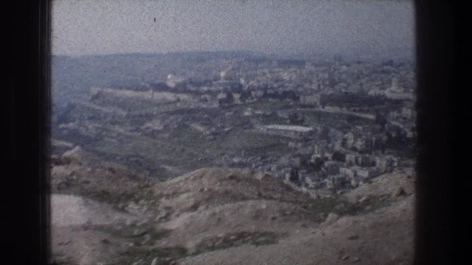 1976 home-movie frame of Jerusalem from an elevated overlook, showing sloped terrain, pale buildings, and a hazy skyline.