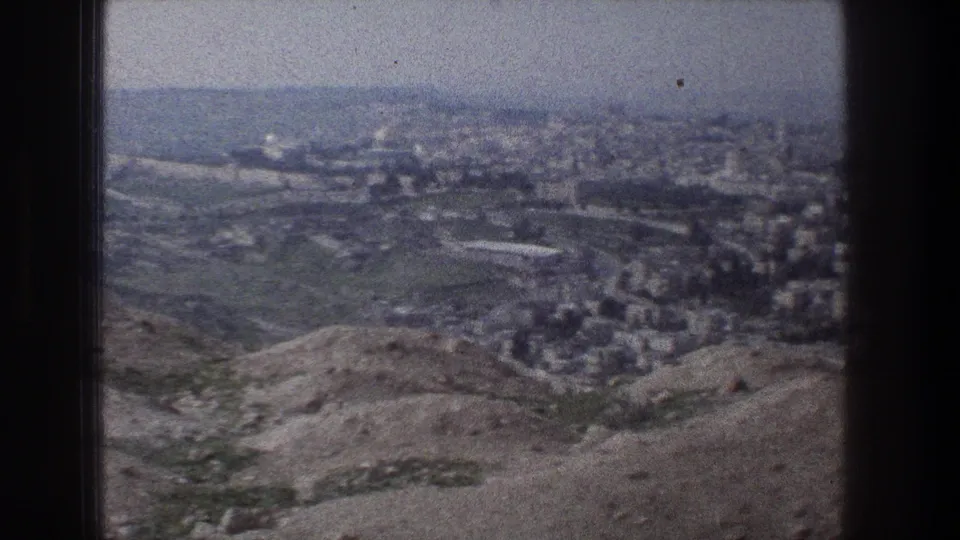 1976 8mm view over Jerusalem from a hillside overlook, with rocky foreground slopes and dense buildings in the distance.