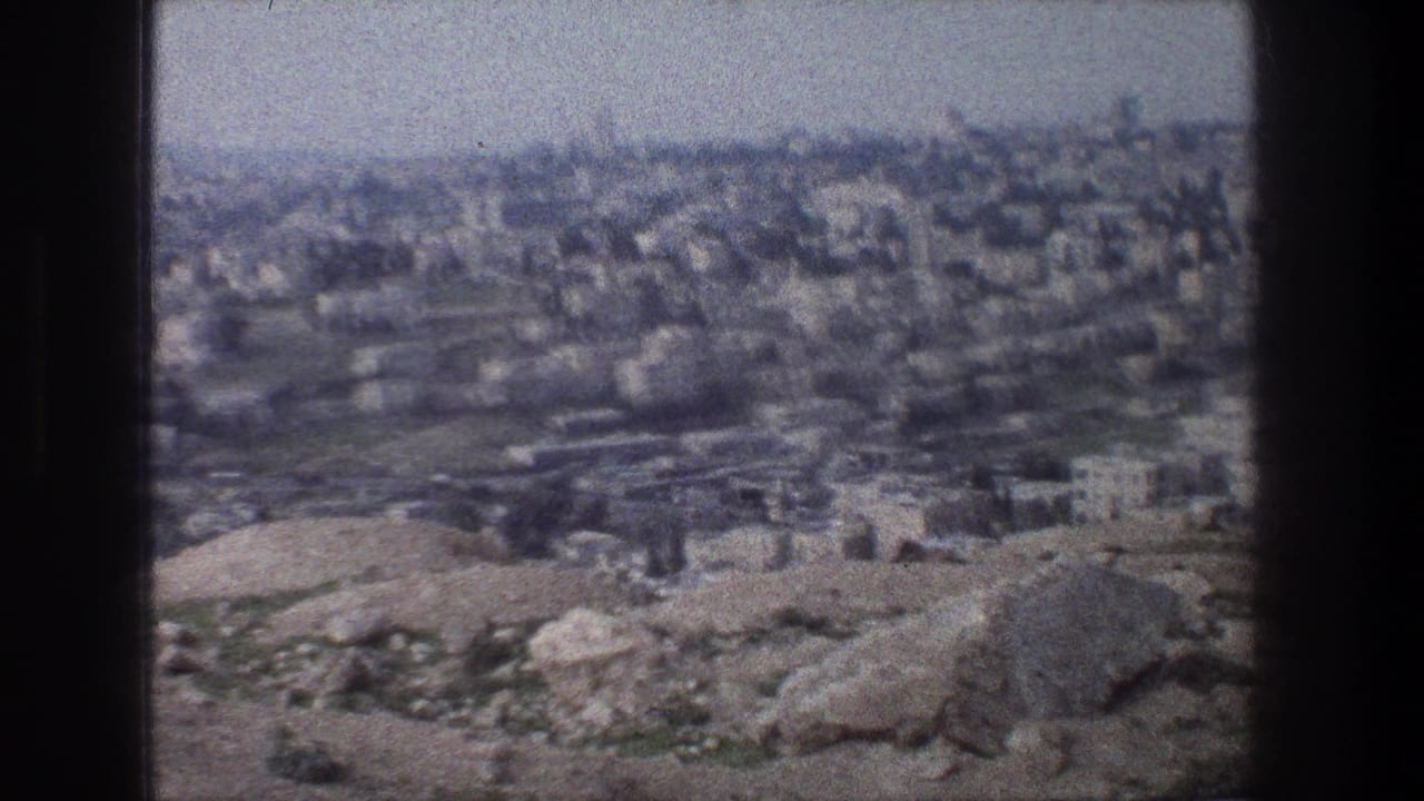 1976 8mm Jerusalem view with pale stone buildings across a hazy hillside and rough rocks in the foreground.