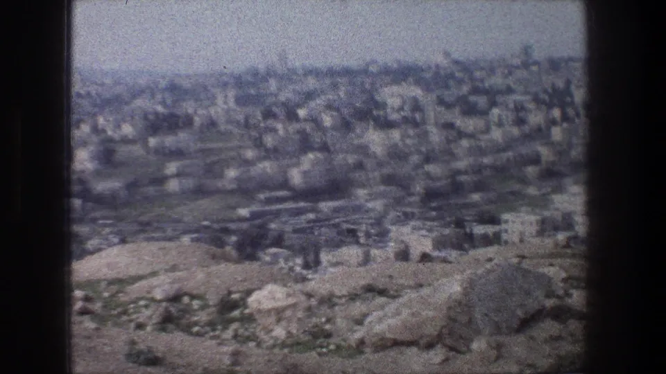 1976 8mm Jerusalem view with pale stone buildings across a hazy hillside and rough rocks in the foreground.