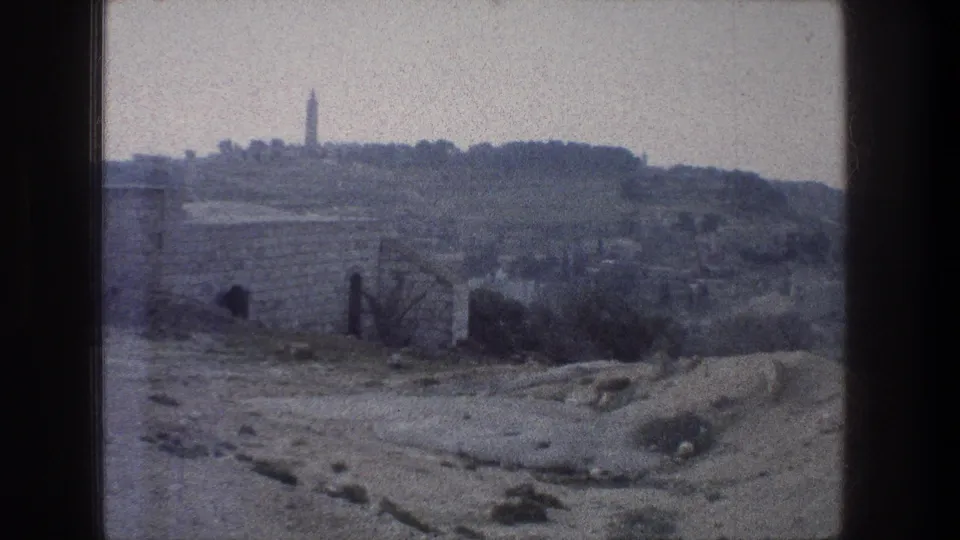 1976 Jerusalem 8mm view toward a hilltop tower or obelisk, with low stone walls, brush, and rough ground in the foreground.