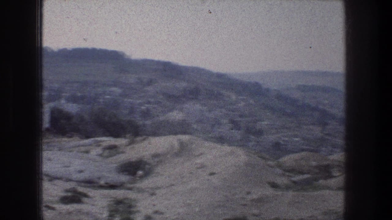 1976 Jerusalem travel-film frame showing pale rocky ground, scattered shrubs, and layered hills under a hazy sky.