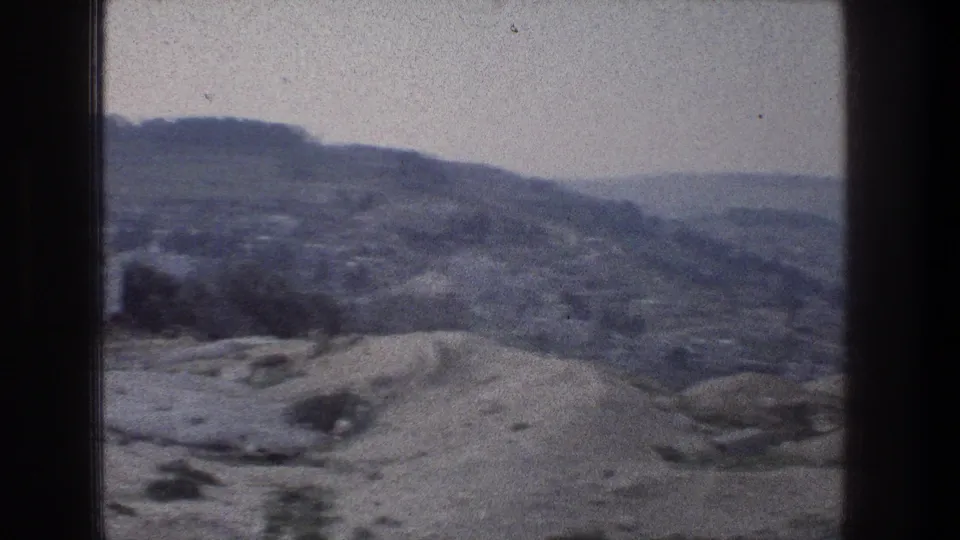 1976 Jerusalem travel-film frame showing pale rocky ground, scattered shrubs, and layered hills under a hazy sky.