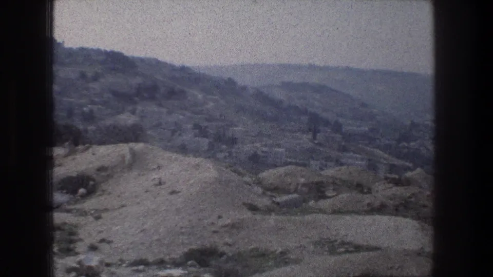1976 8mm frame from Jerusalem showing an elevated view over rocky slopes toward a hazy valley and distant urban forms.