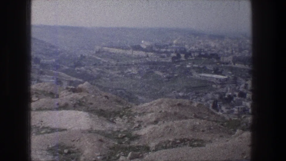 1976 8mm view of Jerusalem from a hilltop, with dry foreground slopes and a hazy distant urban skyline.
