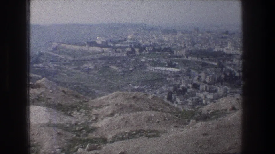 A distant Jerusalem city view holds across dry hills, pale ground, and sparse vegetation in grainy 1970s 8mm color.