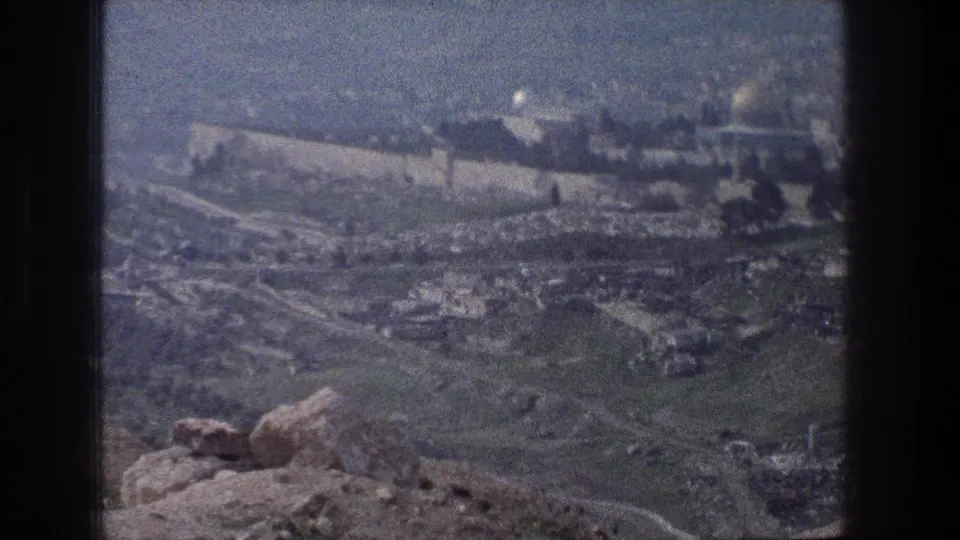 Soft 1976 8mm frame of Jerusalem walls and clustered buildings seen across hillside roads from a rocky overlook.