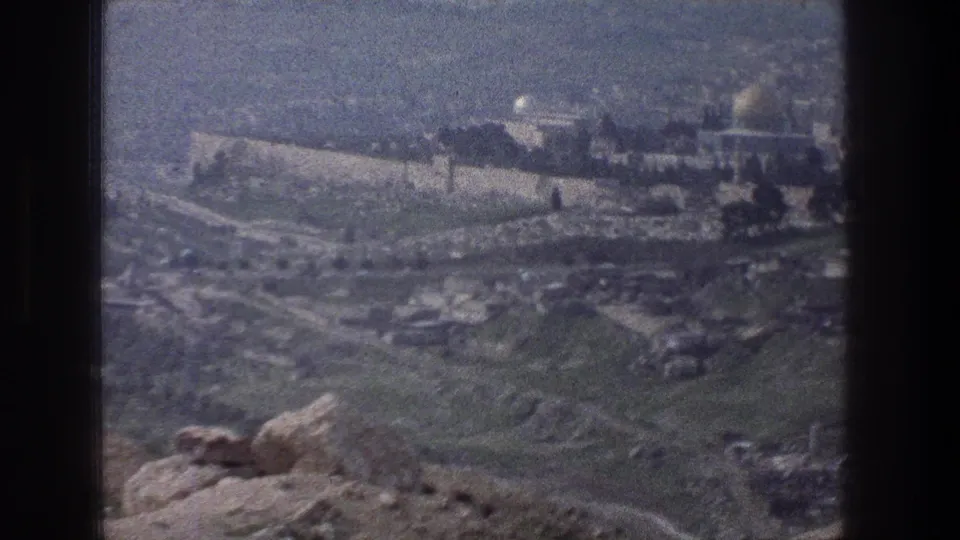 1976 8mm Jerusalem panorama showing tan walls, a distant dome, hillside roads, and rocky ground in the foreground.