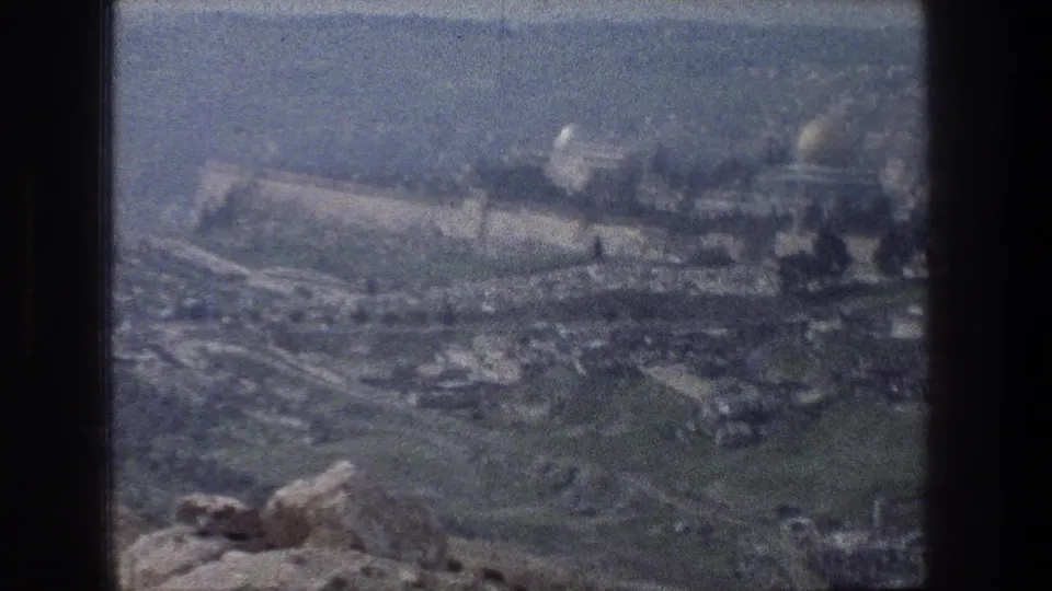 Wide 1976 Jerusalem travel frame showing foreground rocks, the old city wall, distant domes, and soft grain from 8mm film.