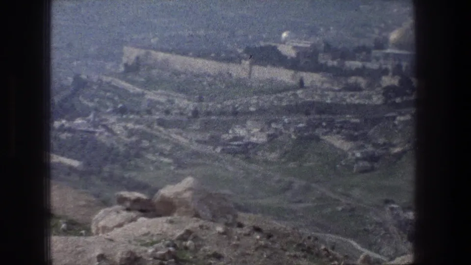 Vintage 8mm Jerusalem overlook with a rocky foreground, hillside paths, stone walls, and a hazy city backdrop.